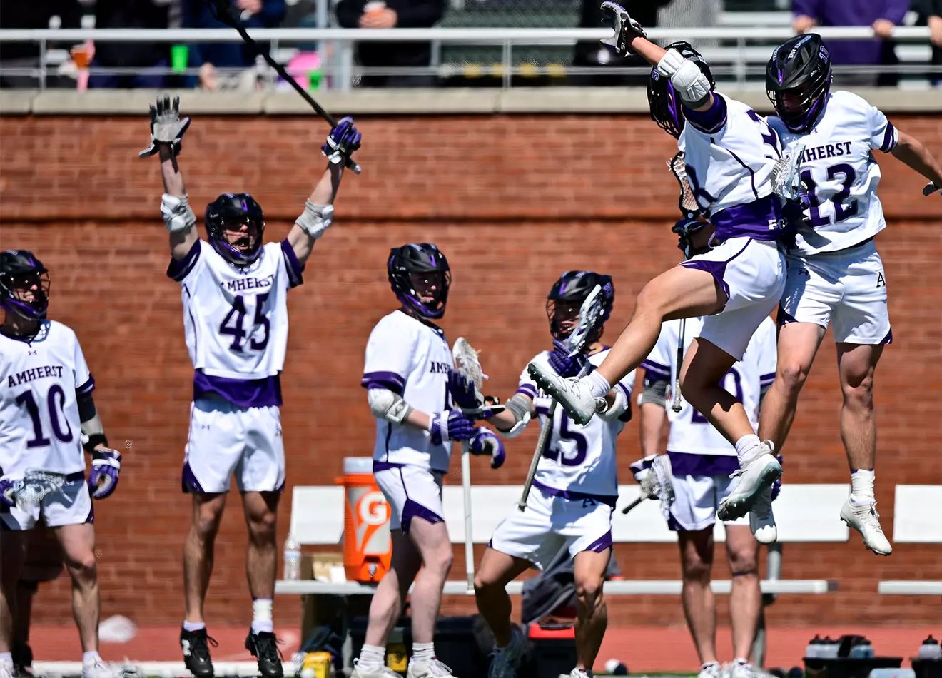 Amherst players celebrating on the field, two leaping together as teammates raise their sticks.